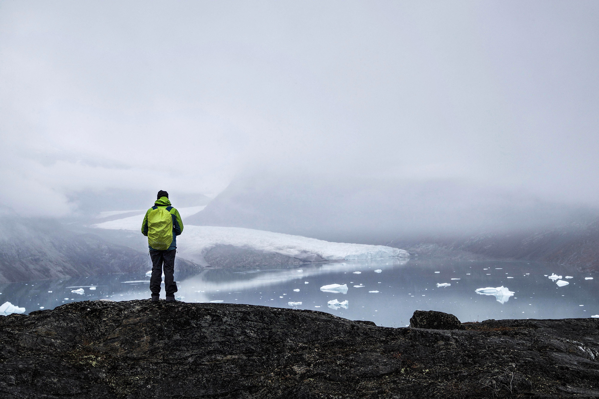 Person i gul jakke står på klippe og ser ud over en tåget isfjord