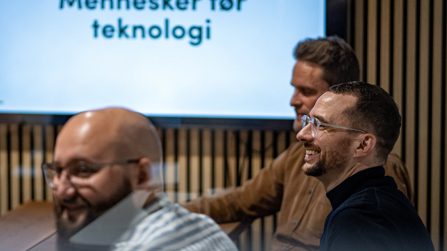 Tre smilende mænd deltager i et møde foran en skærm med teksten