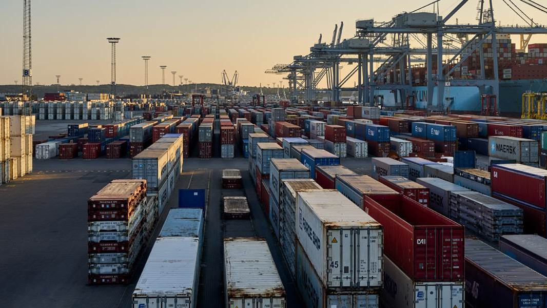 Containeropbevaring på en travl havnekaj med kraner og et skib i baggrunden ved solnedgang