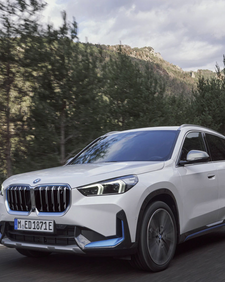 Hvid SUV kører på en vej omgivet af træer og bjerglandskab