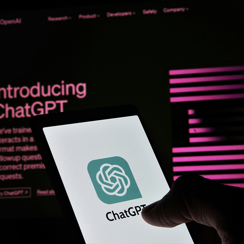 En smartphone viser ChatGPT-logoet, mens introduktion til ChatGPT vises på en computerskærm i baggrunden.