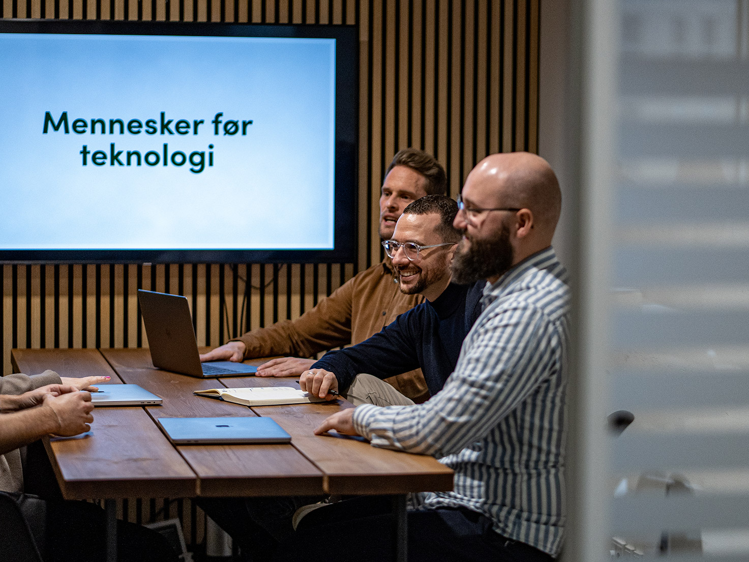 Kollegaer, der smilende diskuterer ved et mødebord foran en skærm med teksten