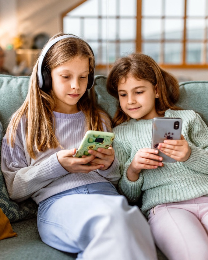To piger sidder på en sofa med smartphones i hænderne, den ene har hovedtelefoner på