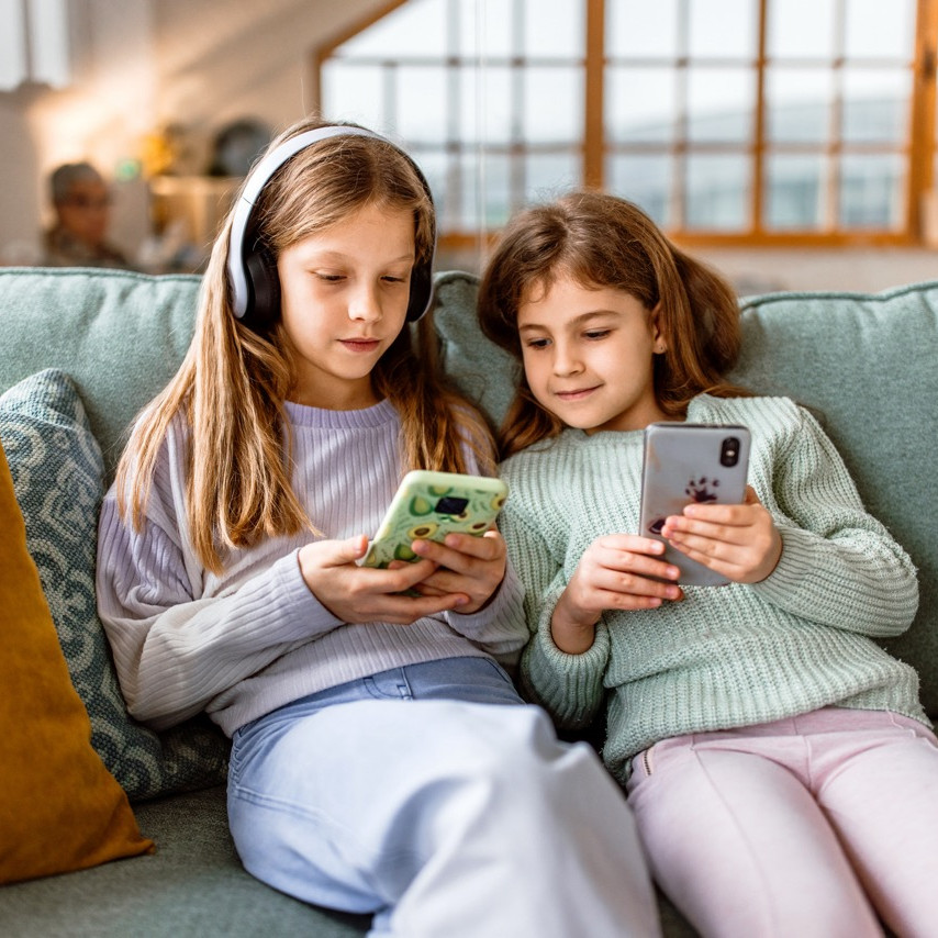 To piger sidder på en sofa med smartphones i hænderne, den ene har hovedtelefoner på